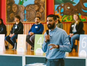 A man holding a microphone stands in front of a panel of people in a New Zealand school hall