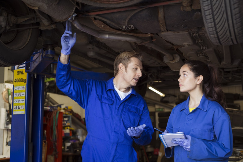 Apprentice mechanic holding a notepad looks at her trainer as he points to the muffler system of a car on a hoist above them.