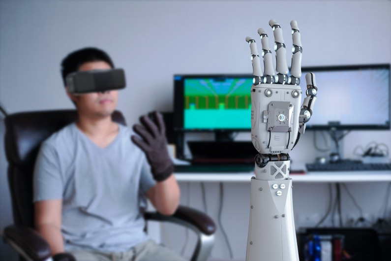 In the foreground is a robotic hand. In the background a man sits with a virtual reality headset on and wearing a glove that controls the robotic hand.