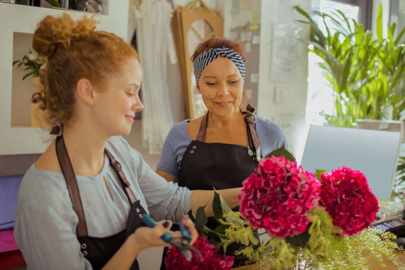 A young and older woman wearing casual clothes and aprons stand in a florist shop. The younger one is working with a big bunch of red flowers while the other one looks on
