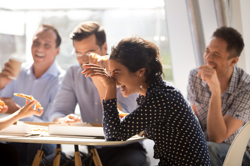 A woman laughs while eating pizza with her office co-workers