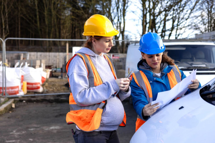 Two females wearing hi-vis and helmets, standing on a housing development plot and looking at the plans on a piece of paper . One of the women is 7 months pregnant.