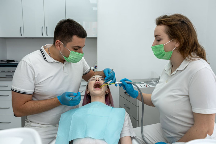 A male dentist works on a child's teeth while the dental assistant holds a suction tube in the mouth