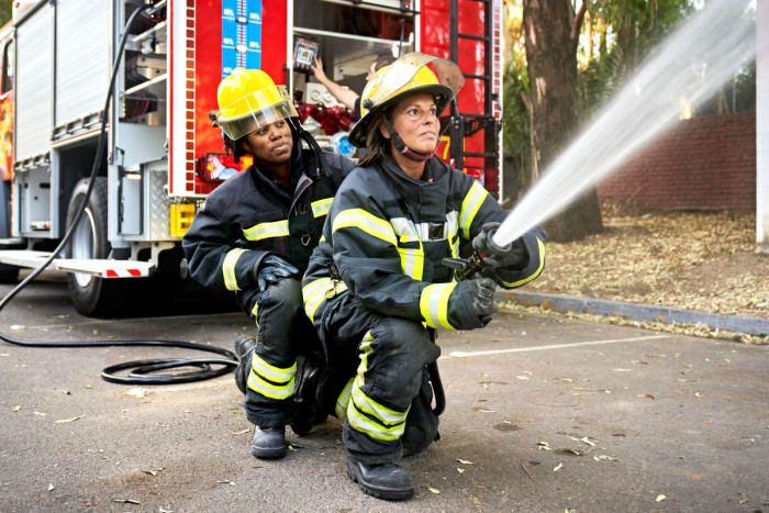 Two female firefighters in full uniform and protective helmets point a fire hose at a fire