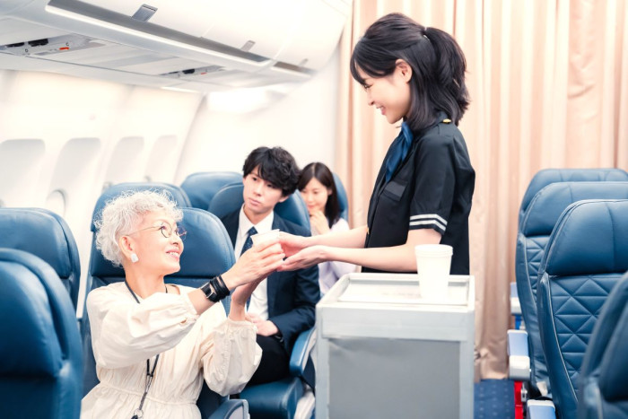 A flight attendant in an aeroplane cabin hands a drink to a woman sitting in an aisle seat