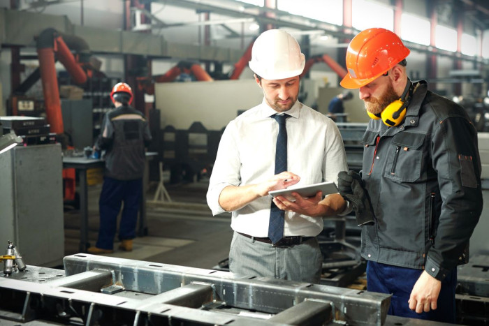 Two men in hard hats, one wearing overalls and earmuffs and the other in office clothes stand over a machine and ook at a digital tablet whille standing in a factory