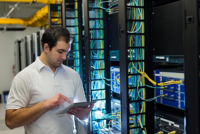 A man in a server room with many cables looking at a tablet 