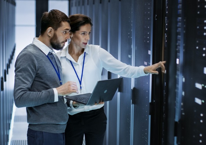 A man holding a laptop talks to a woman who is pointing to a server in a computer server room 